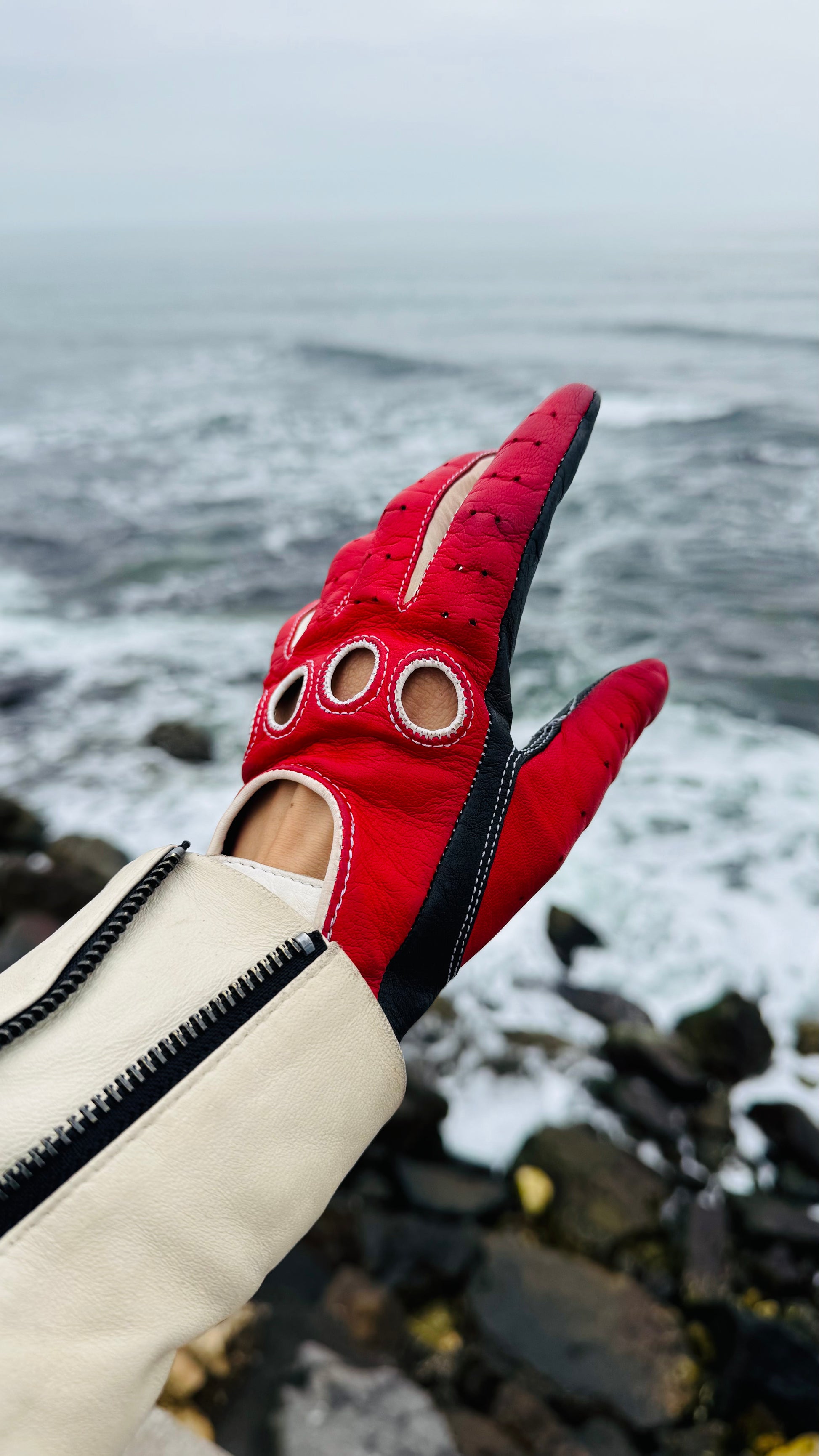 Poseidon tricolour driving glove
A tricolour leather driving glove with black trim and cream baguyettes, set against a blurred rocky ocean background.