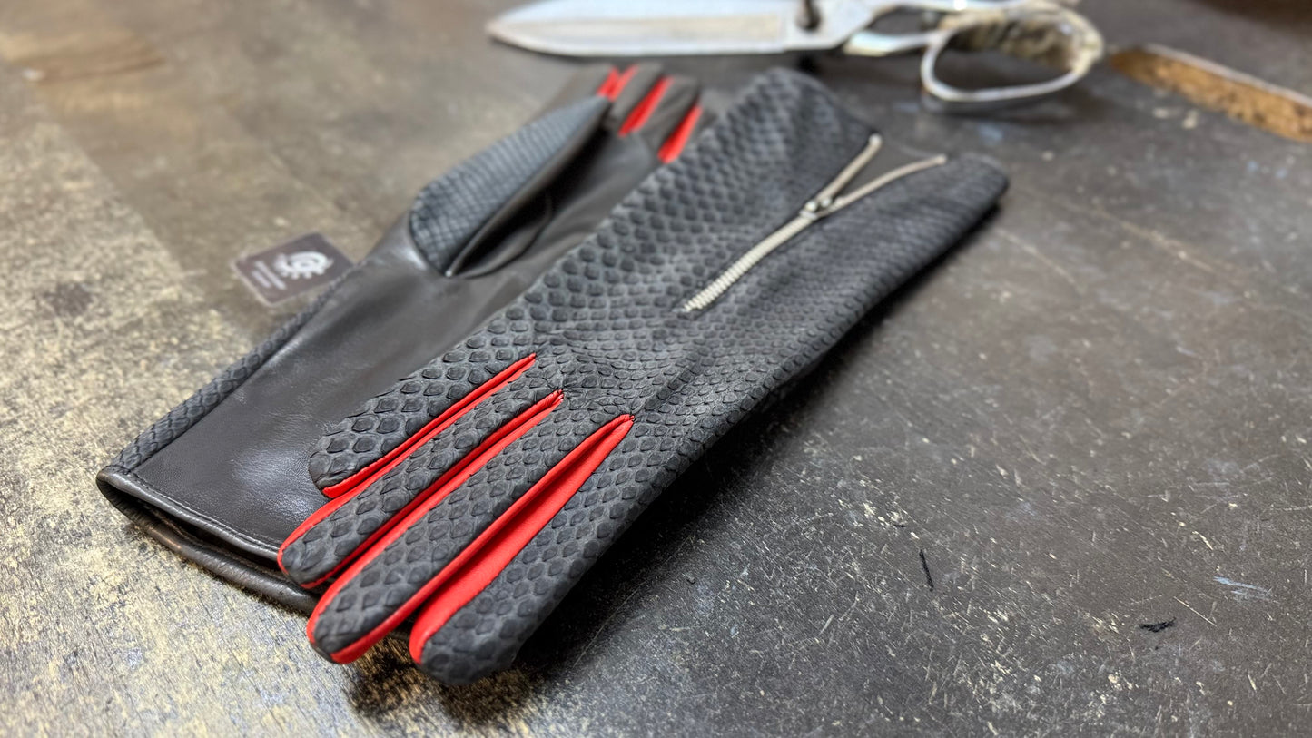 A black glove with red fourchettes between the fingers, lying with the python side up beside the other glove with the lambskin side up. In the background, a pair of scissors to illustrate they are handcrafted.